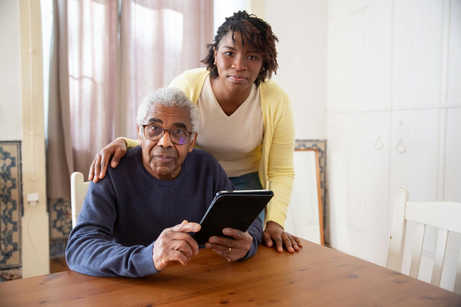 Adult woman supports elderly man using tablet at home.
