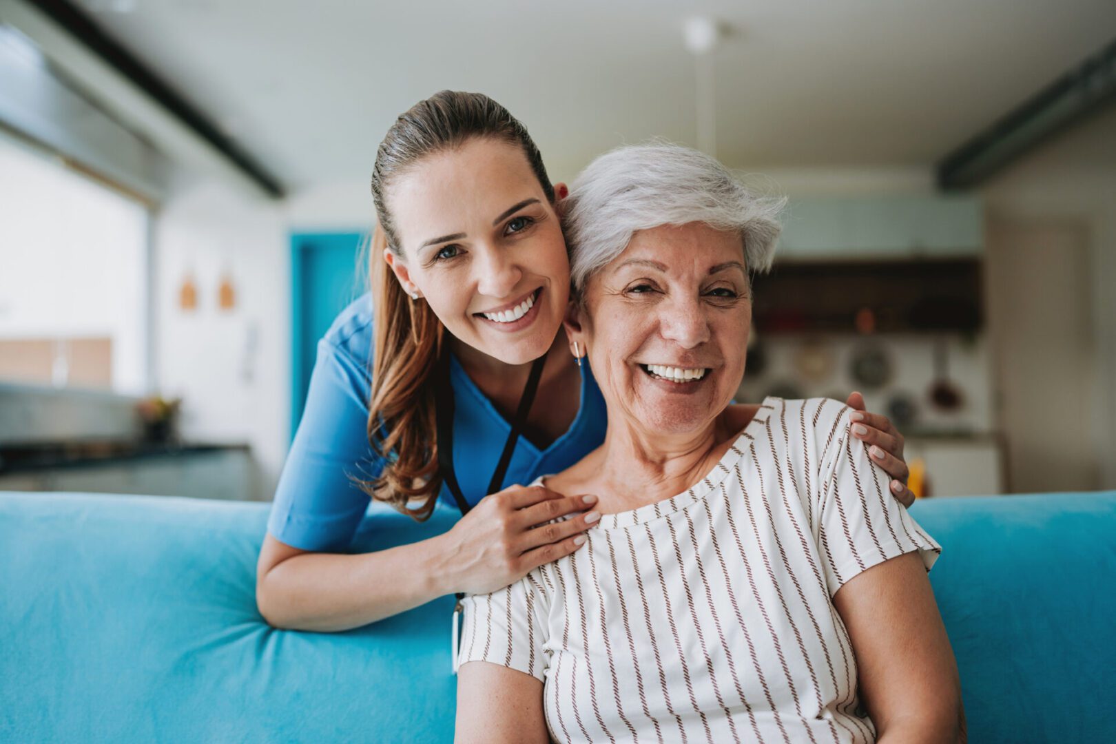 A young caregiver warmly embraces an elderly woman, both smiling happily indoors.